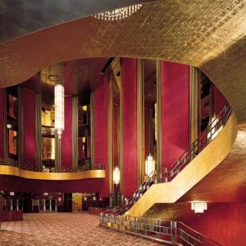 Interior of the Radio City Music Hall Lobby in New York City, New York featuring an ornate curved ceiling with gold leaf detailing, elegant red draperies, and classic hanging light fixtures.