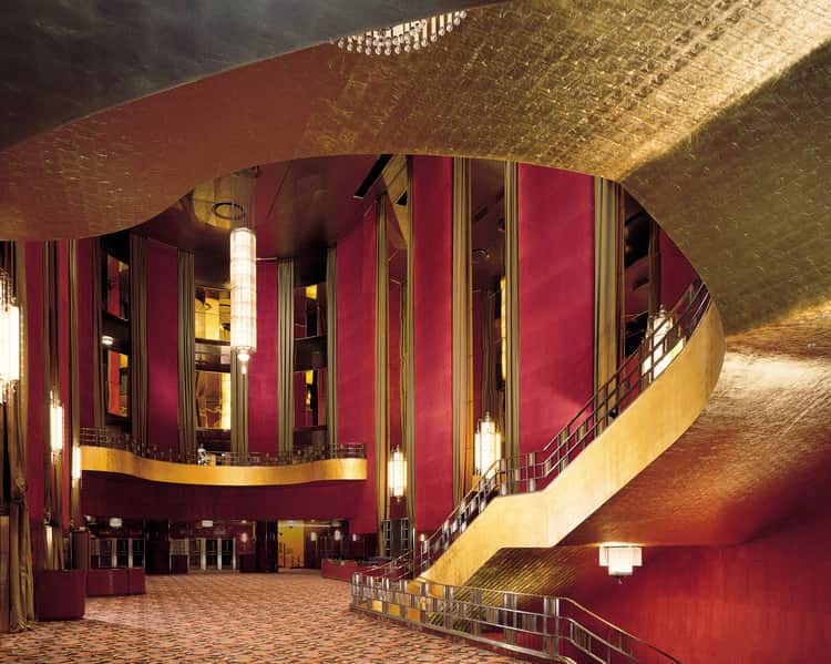 Interior of the Radio City Music Hall Lobby in New York City, New York featuring an ornate curved ceiling with gold leaf detailing, elegant red draperies, and classic hanging light fixtures.